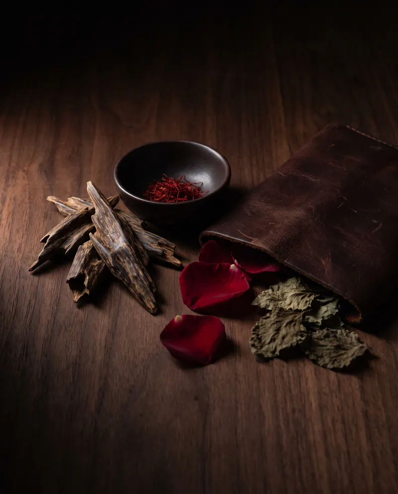 The perfumer’s table: oud wood chips, saffron in a small bowl, rose petals, a leather pouch and patchouli leaves